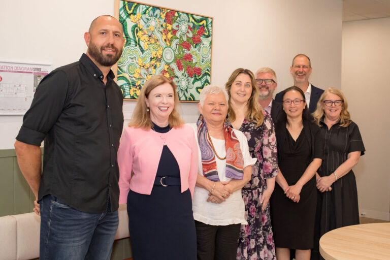 Group of people standing in front of an Aboriginal Artwork smiling.