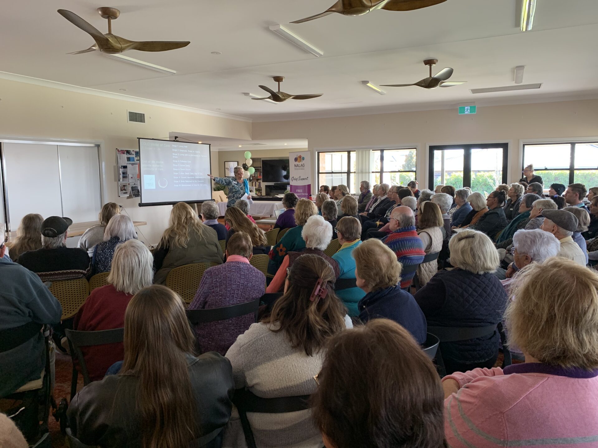 A room full of people looking at a presenter in front of a projector screen.