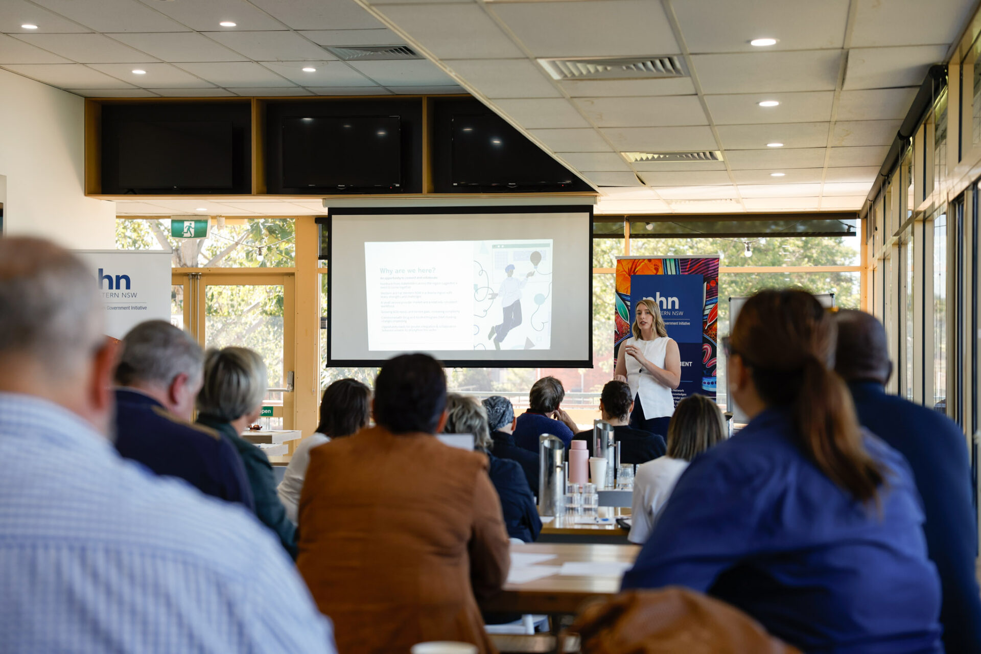 Photograph of a presentation in a conference room with attendees seated facing a screen displaying a slide with text and graphics. Presenter stands near screen, with banners on either side, and natural light coming through large windows on the right.