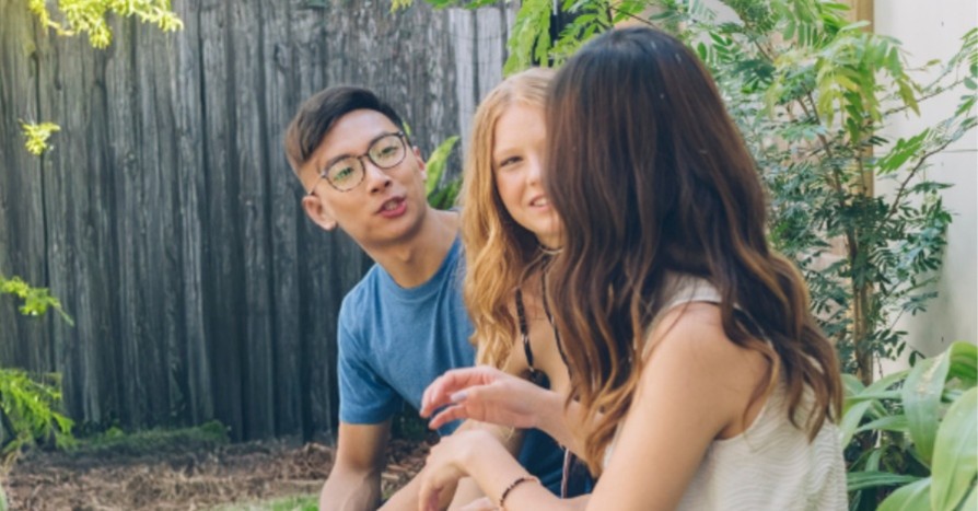 Photograph of three people sitting outdoors in a garden or backyard setting, engaged in conversation. Surrounding elements include wooden fencing, green plants, and natural sunlight, creating a relaxed and casual atmosphere.