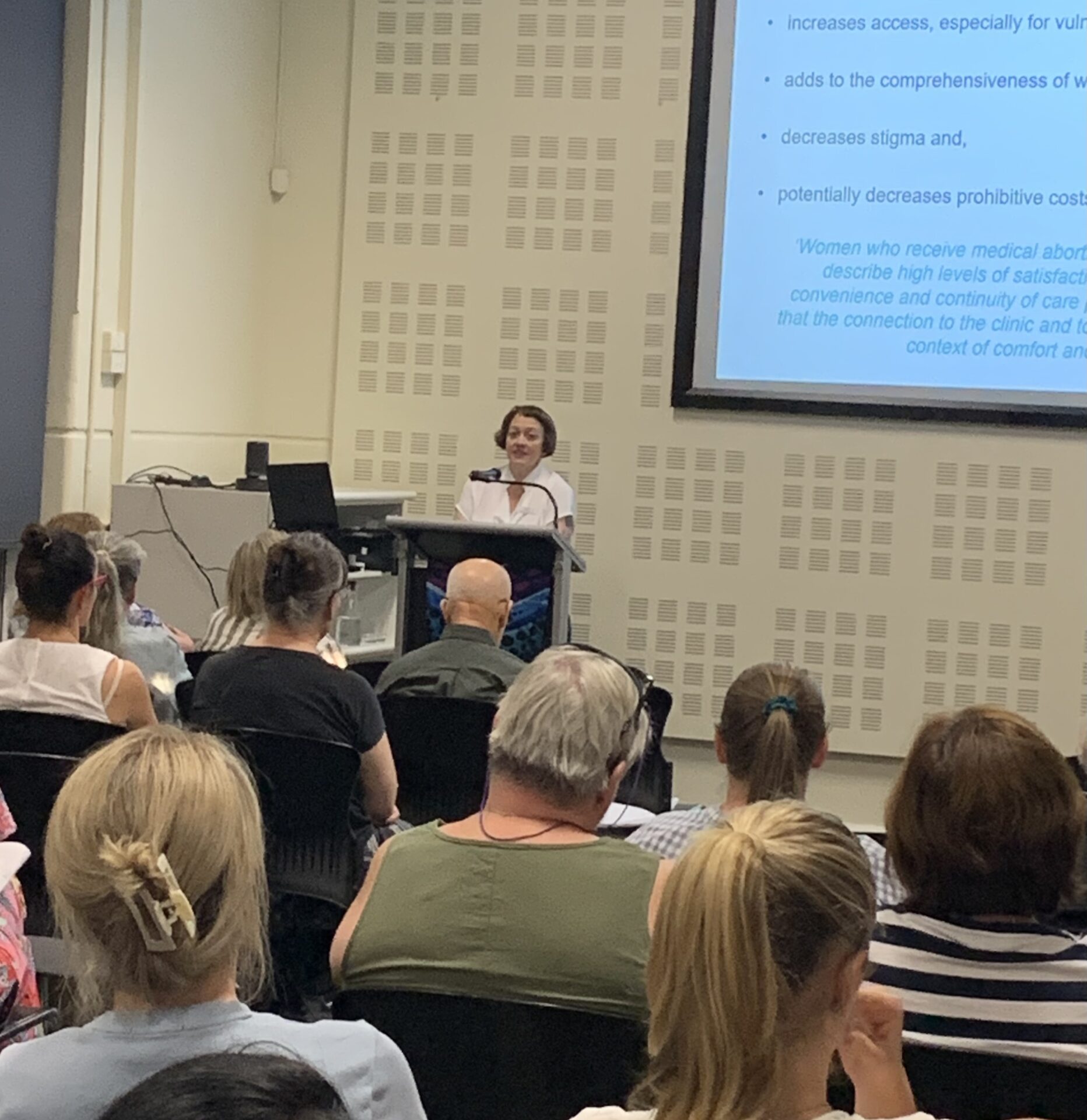 Photograph of a conference or seminar setting with an audience facing a speaker at a podium and a projected presentation slide on a wall. Slide content includes bullet points discussing benefits such as increased access, decreased stigma, and potential cost reductions related to medical services, with highlighted text about women's satisfaction and care continuity.