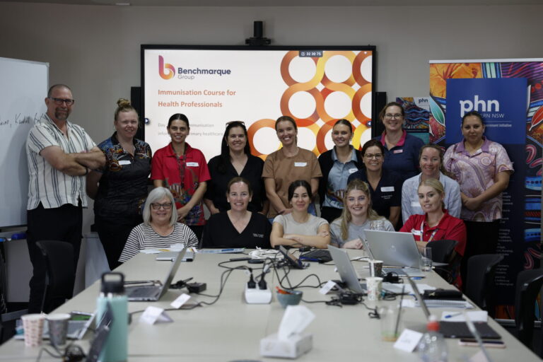 Photograph of a group of health professionals posing in a conference room with laptops and documents on a table. A presentation slide on a screen behind them displays "Immunisation Course for Health Professionals" by Benchmarque, indicating a training or educational session.