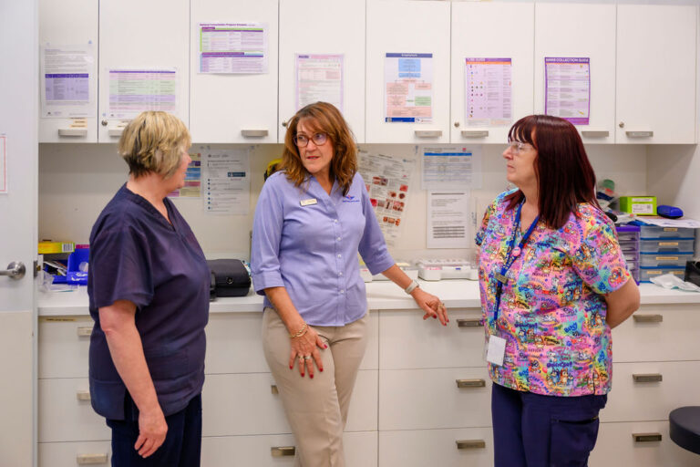 Three women standing and talking, there are cupboards behind them with posters placed on cupboard doors