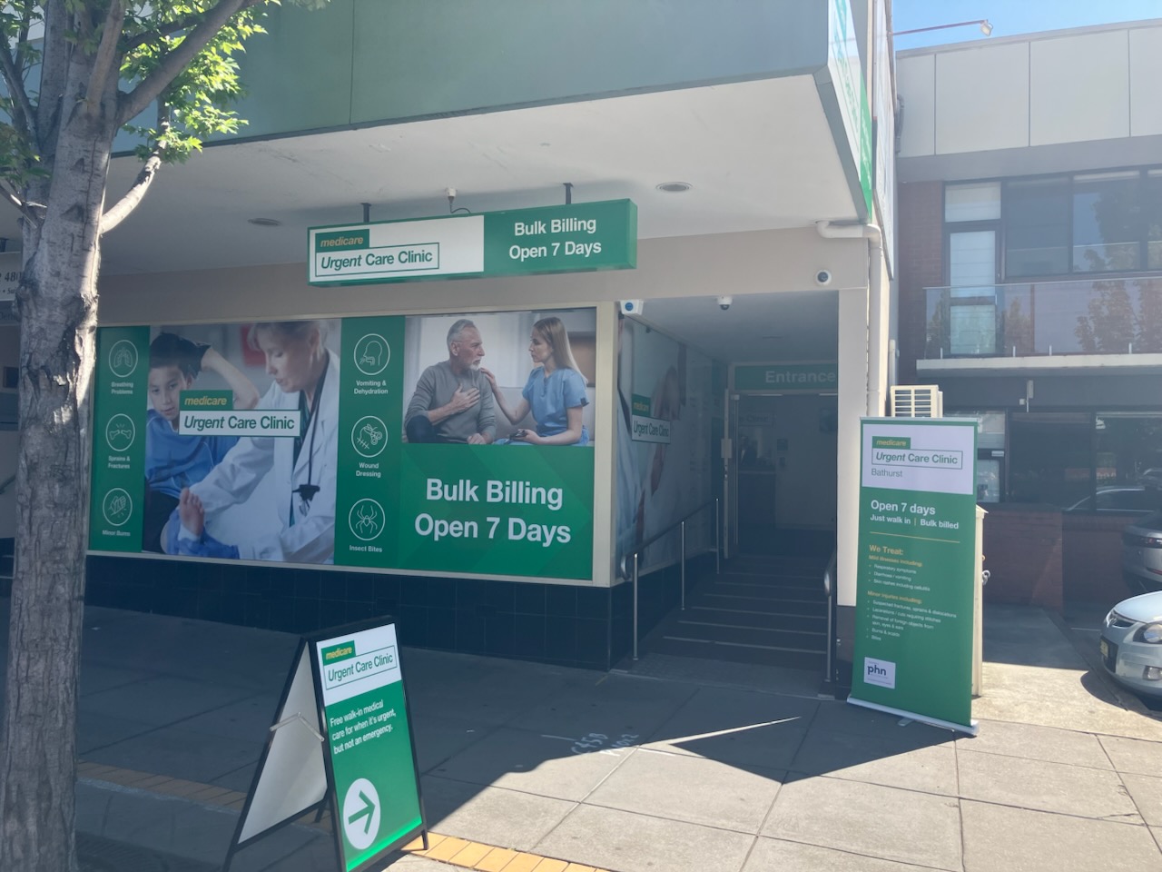 Photograph of an urgent care clinic storefront with multiple green and white signs advertising bulk billing and seven-day availability. Signs include images of medical professionals and patients, directional arrows, and clinic hours, highlighting accessibility and affordability of healthcare services.