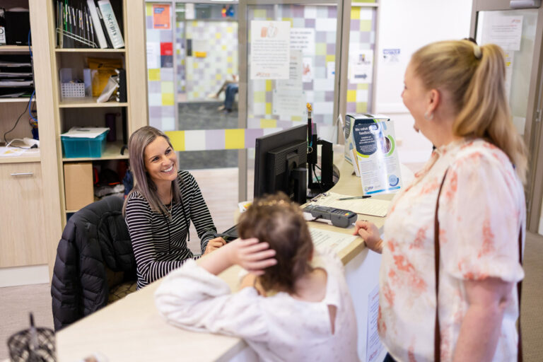 Woman sitting behind a desk at a medical centre smiling at a woman and child on the other side of the desk
